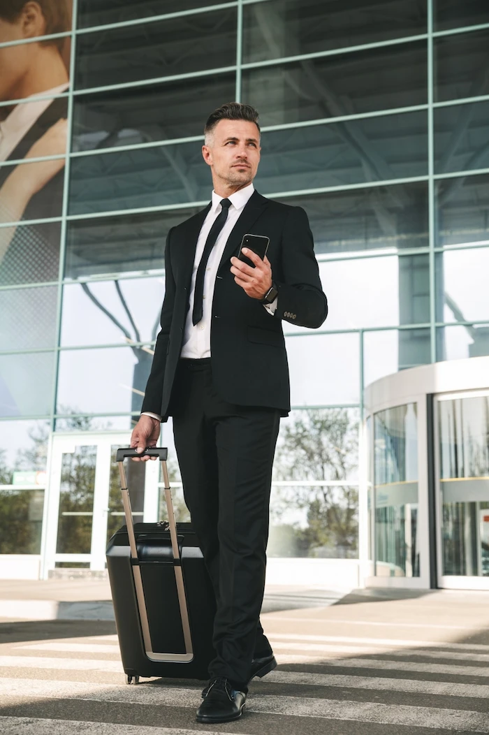 Executive with rolling luggage and smartphone walking outside an airport terminal for car service pickup